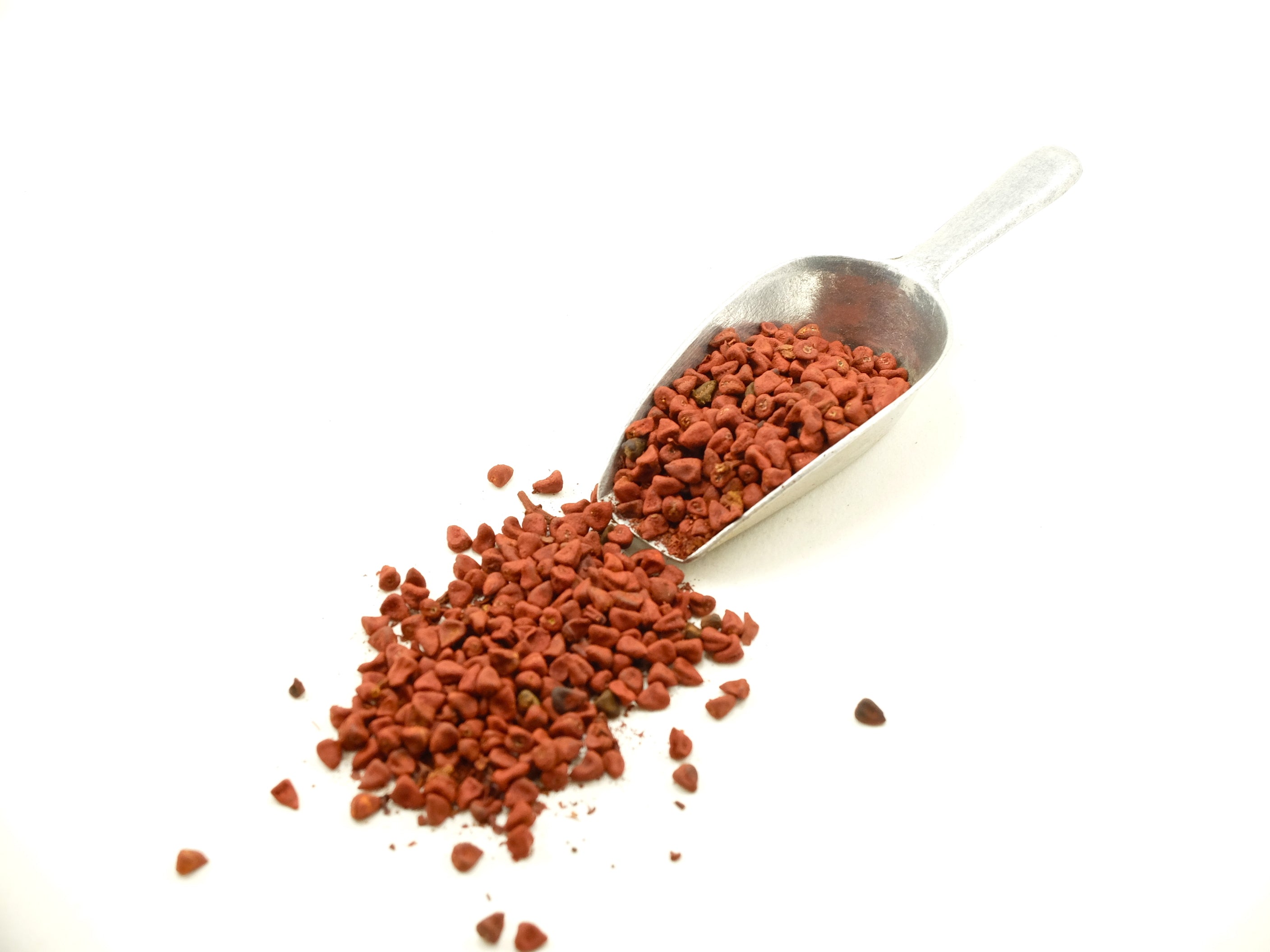 A metallic scoop filled with small reddish-brown annatto seeds spilling onto a white surface